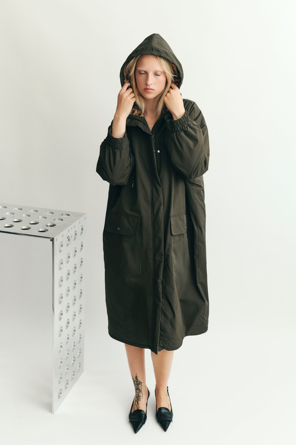 Model holding the hood of an oversized dark olive parka with gathered sleeves, large front pockets and brown patterned collar trim.