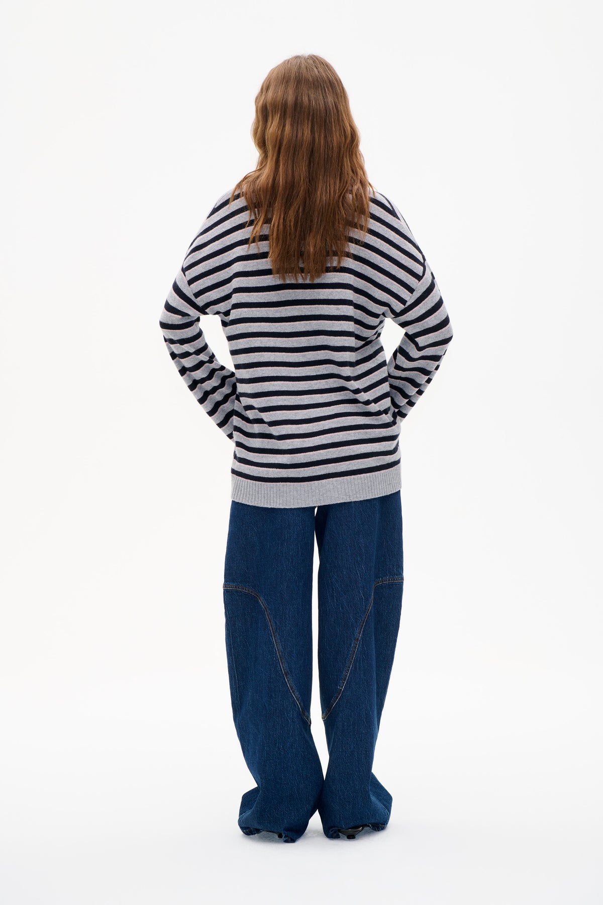 Back view of a model wearing a grey knit sweater with navy and pale pink horizontal stripes, paired with wide-leg dark blue jeans.