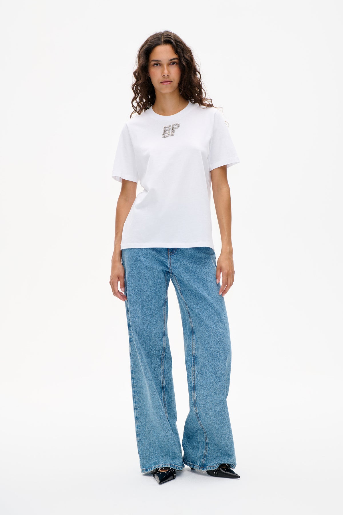 Front view of a model wearing a white short-sleeved T-shirt with a jeweled letter emblem, blue wide-leg jeans and black pointed shoes.