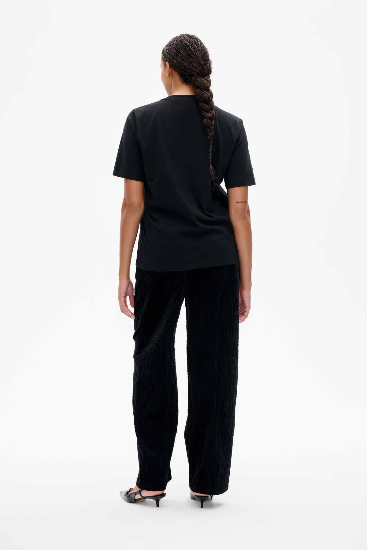 Rückenansicht eines Models, das ein schlichtes schwarzes Kurzarm-T-Shirt und eine schwarze, strukturierte, verkürzte Hose trägt.
