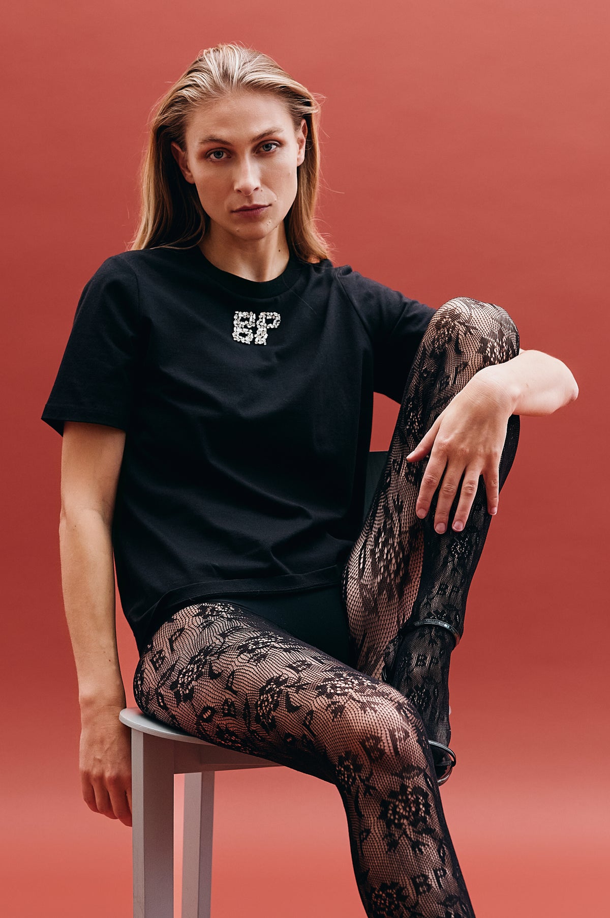 Model sitzt vor korallenfarbenem Hintergrund und trägt ein schwarzes, kurzärmeliges Top mit funkelndem Brustmotiv sowie eine schwarze Strumpfhose mit Spitzenmuster.