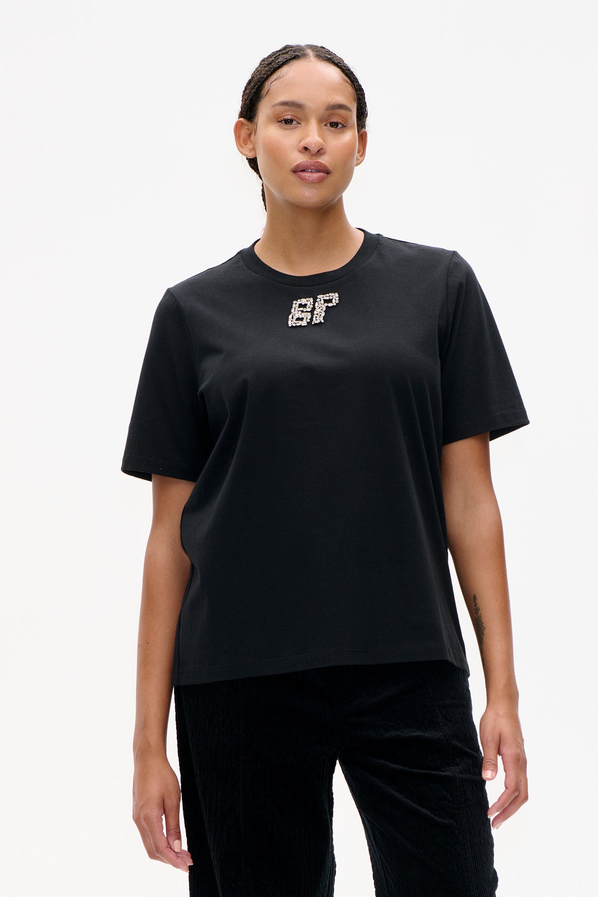 Model wearing a black short-sleeve crew-neck T-shirt with a small silver jeweled chest motif and black ribbed trousers.