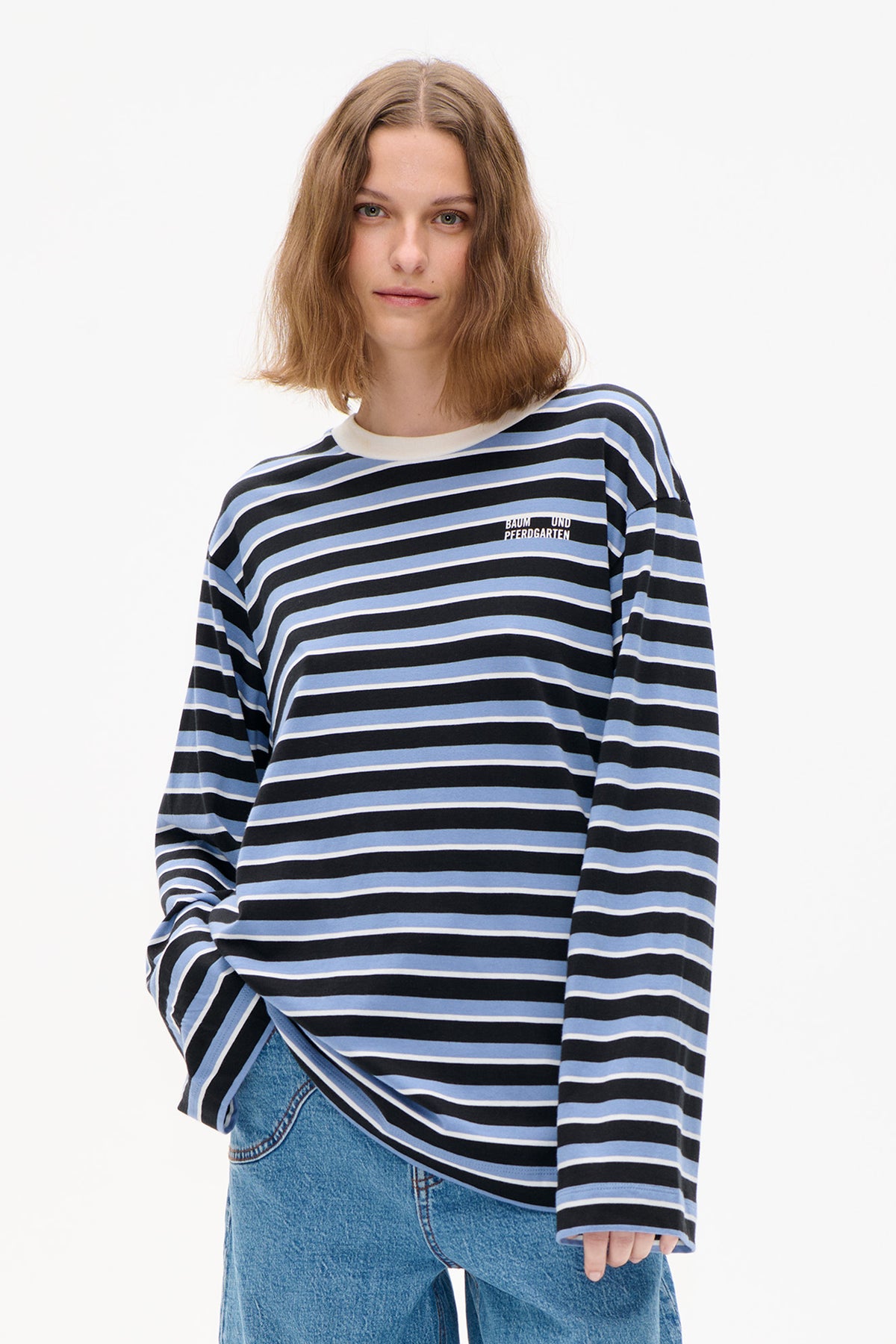 Frontansicht eines Models, das ein langärmliges Top mit schwarzen, hellblauen und weißen Streifen und kleinem Brustlogo trägt, kombiniert mit einer blauen Jeans.