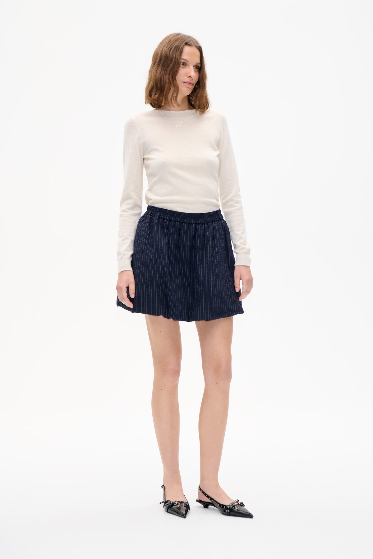 Das Model trägt ein elfenbeinfarbenes, langärmliges Rundhals-Top mit kleinem Brustemblem und marineblaue Shorts mit Nadelstreifen vor weißem Studiohintergrund.