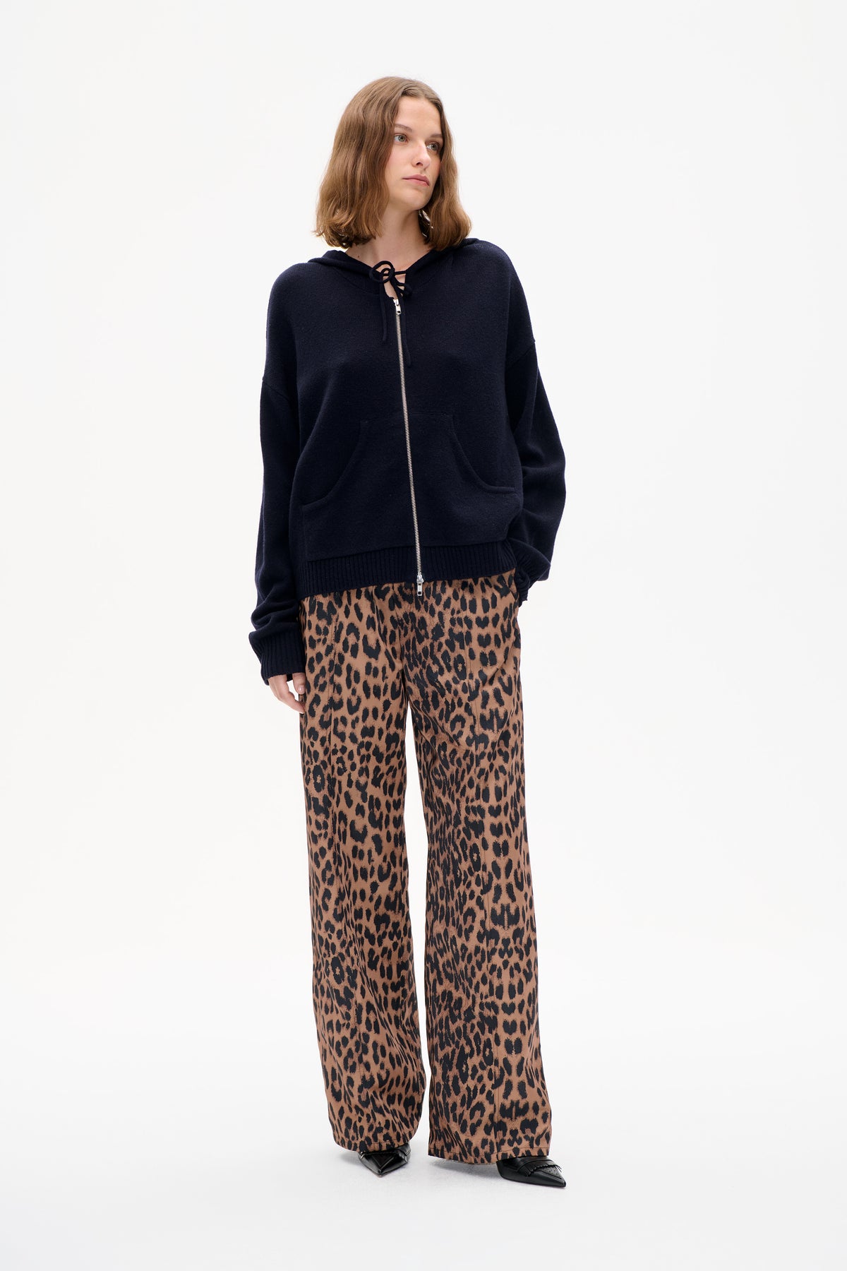 Model wearing a dark navy zip-front hooded sweater with front pockets and brown-and-black leopard-print wide-leg trousers.