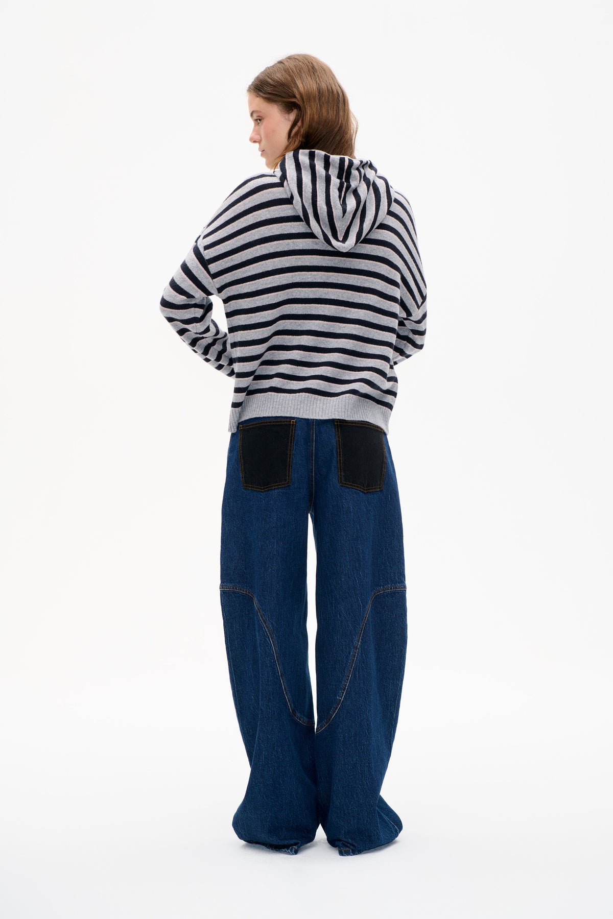 Back view of a model wearing a grey and navy striped hooded knit sweater with wide-leg dark blue jeans and patch pockets.