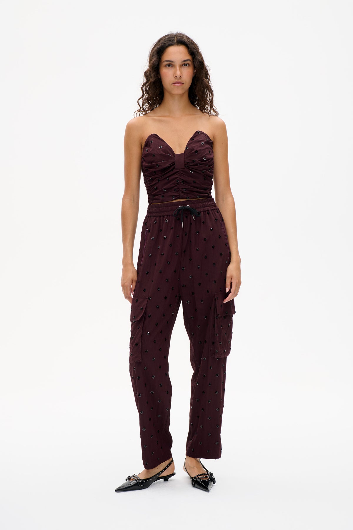 Front view of a model wearing a burgundy strapless ruched top and matching drawstring trousers with scattered black floral embellishments.