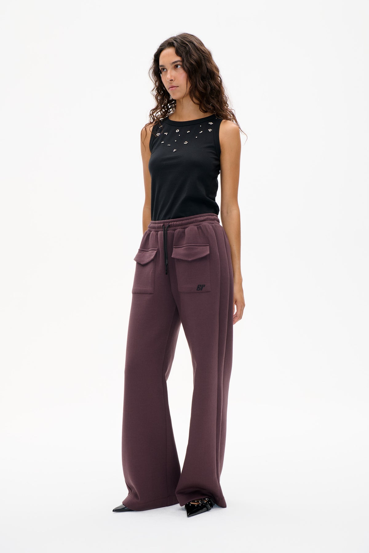 Model trägt ein schwarzes, ärmelloses Top mit vereinzelten silbernen Verzierungen und eine mauvefarbene, weit geschnittene Hose mit Kordelzugbund und aufgesetzten Taschen.