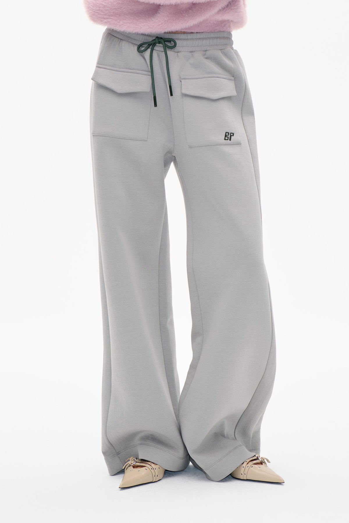 Vorderdetail einer hellgrauen, weit geschnittenen Sweatpants mit grünem Kordelzug, Klappentaschen und kleinem BP-Logo, kombiniert mit einem rosa flauschigen Top.