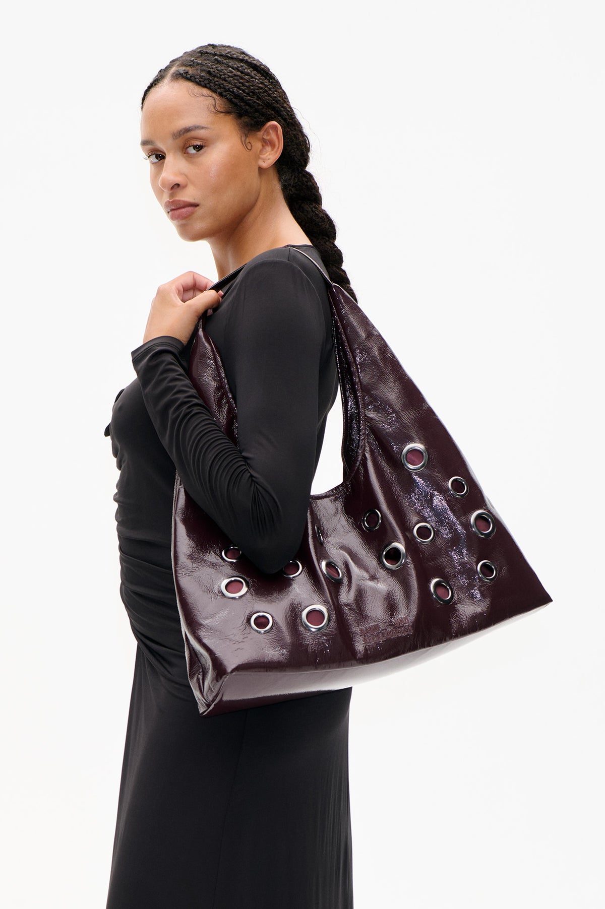 Model in a black outfit carries a glossy dark burgundy tote bag with silver-tone eyelets.