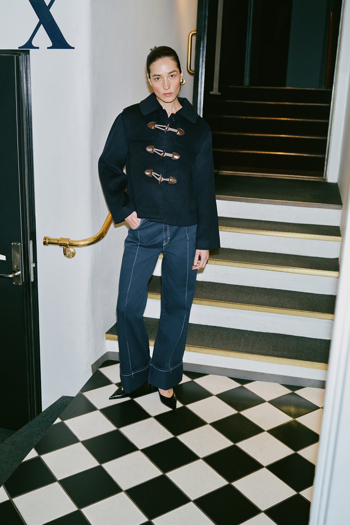Model wearing a dark navy coat with brown toggle fastenings and flap pockets, paired with blue jeans on a tiled staircase.
