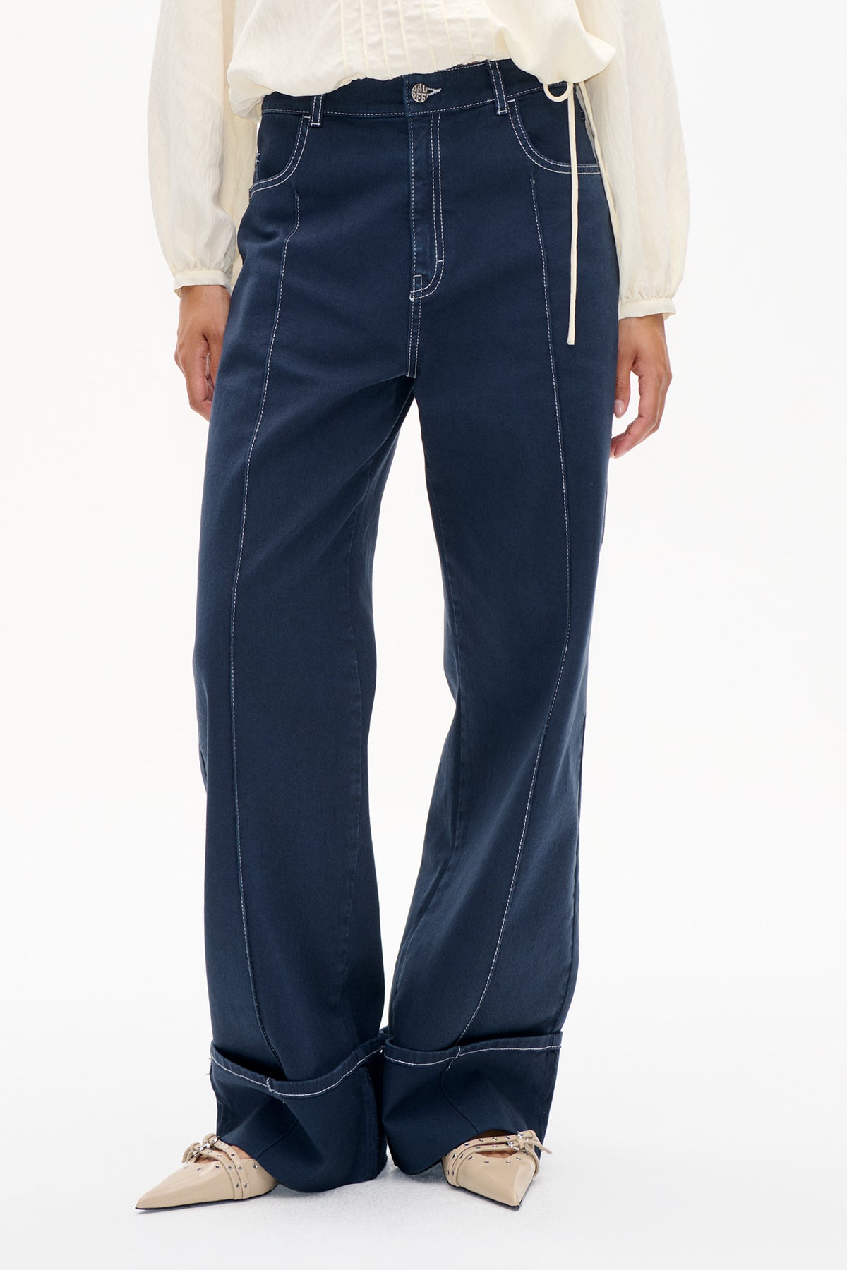 Detail der Vorderseite einer dunkelindigo-Jeans mit markanter weißer Naht, langem Vorderteil und aufgekrempelten Säumen, kombiniert mit einer durchsichtigen elfenbeinfarbenen Bluse.
