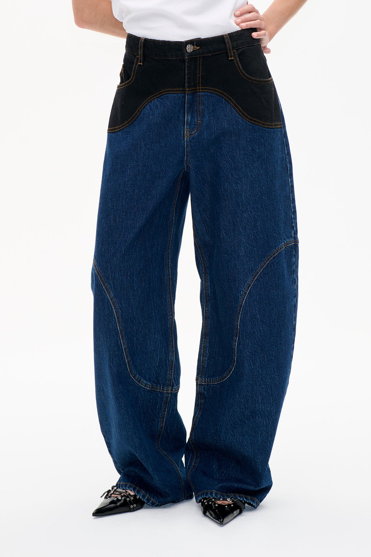 Detail der Vorderseite einer weit geschnittenen blauen Jeans mit schwarzen, geschwungenen Hüftbesätzen, goldfarbenen Kontrastnähten und Knopfverschluss.