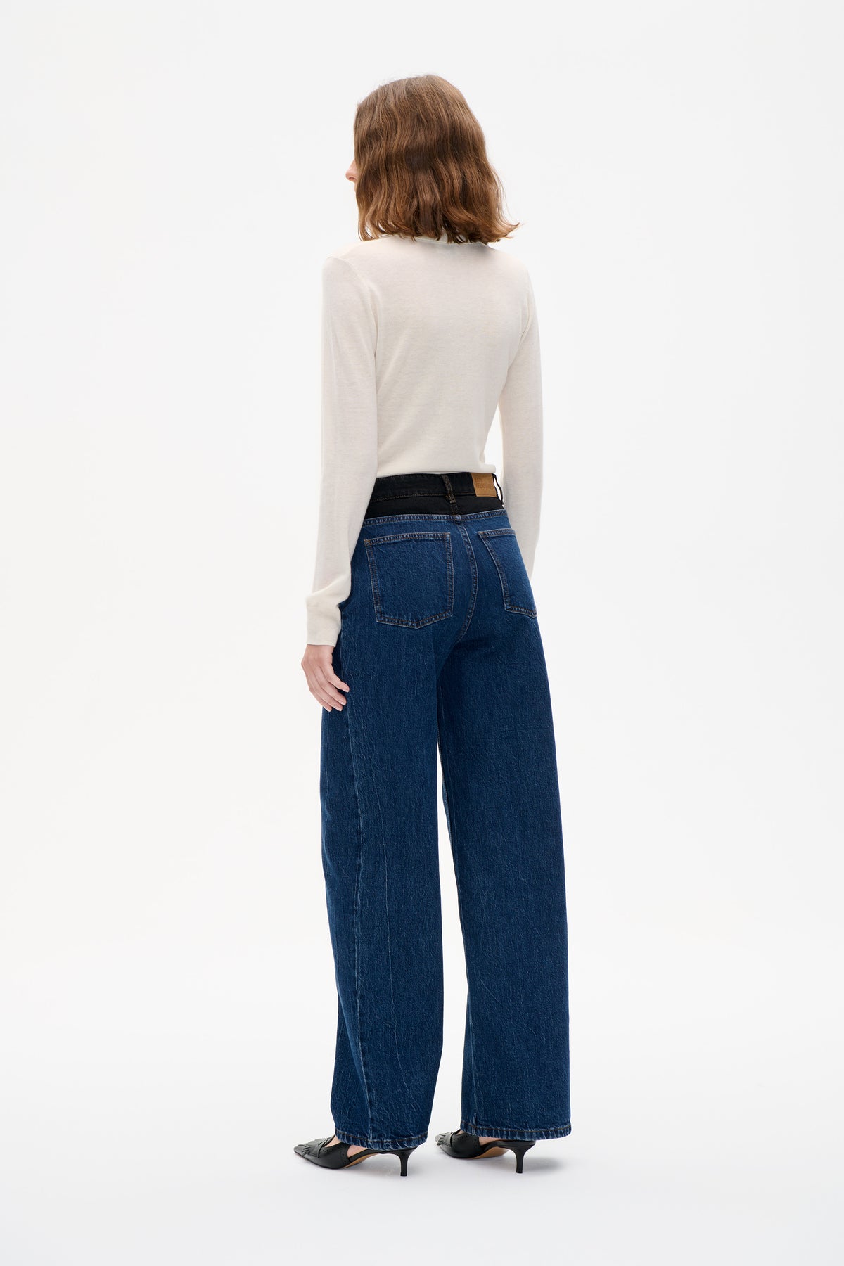 Rückenansicht eines Models, das dunkelblaue Jeans mit weitem Bein, Kontrastnähten und ein cremefarbenes Langarmtop trägt.
