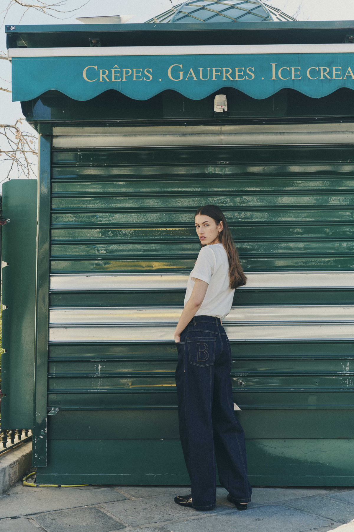 A model looks back beside a green storefront, wearing a white short-sleeve T-shirt and dark wide-leg jeans with outlined B stitching on the back pocket.
