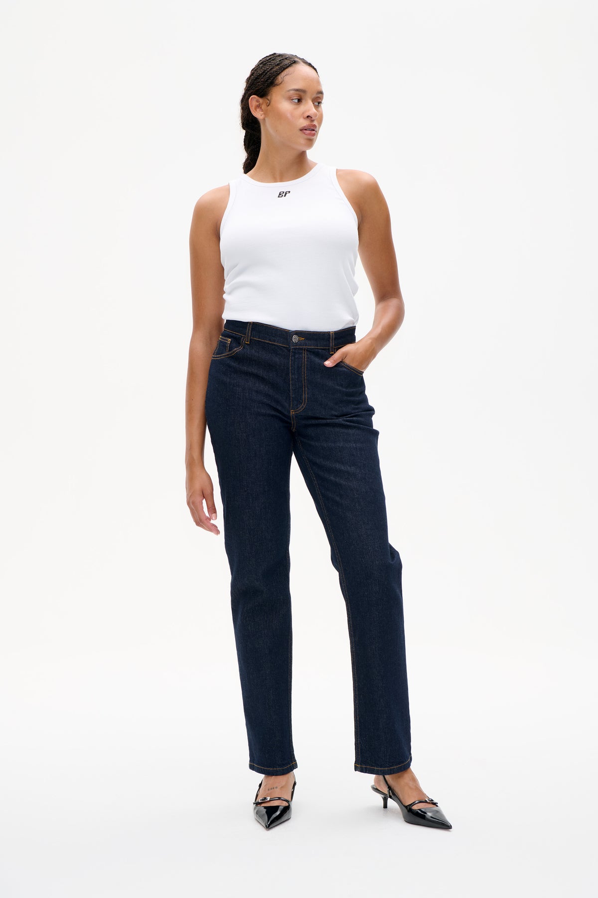Model draagt een witte, mouwloze ribgebreide top met klein borstlogo en donker indigo jeans met rechte pijpen, gezien van voren.