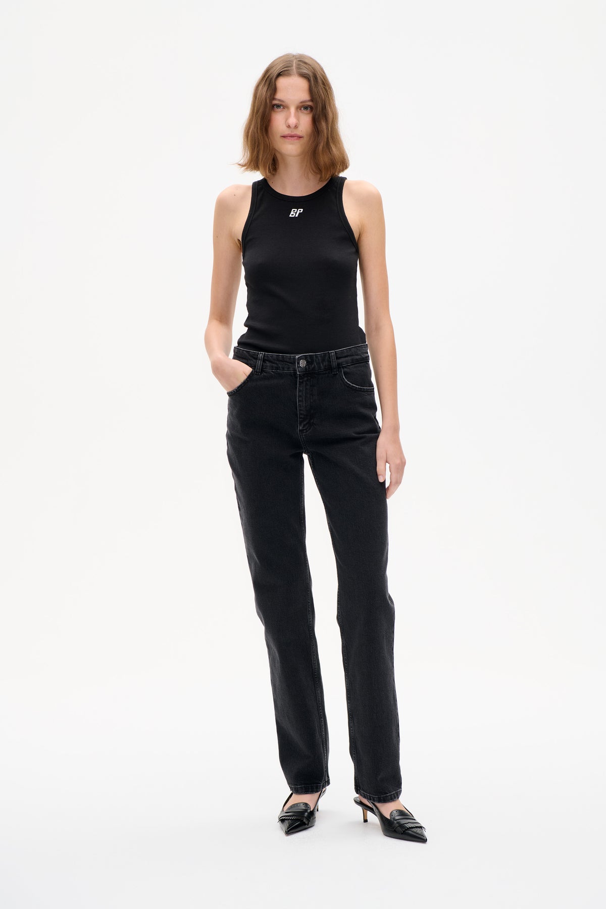 Vooraanzicht van een model in een zwarte mouwloze geribde top met klein wit BP-logo op de borst en donkere jeans met rechte pijpen.