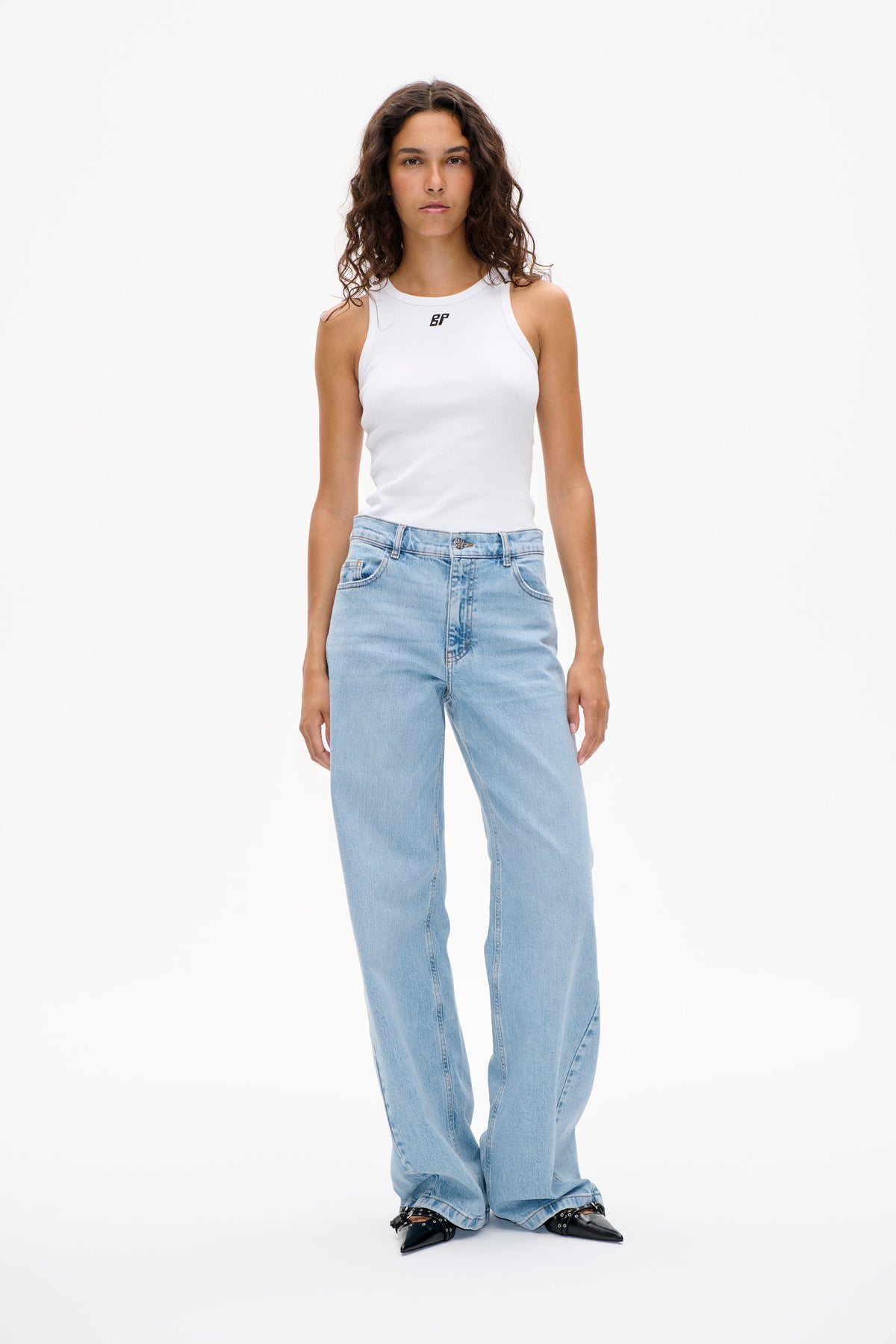 Front studio view of a model wearing a white sleeveless ribbed top with a small chest logo, light blue wide-leg jeans and black pointed shoes.