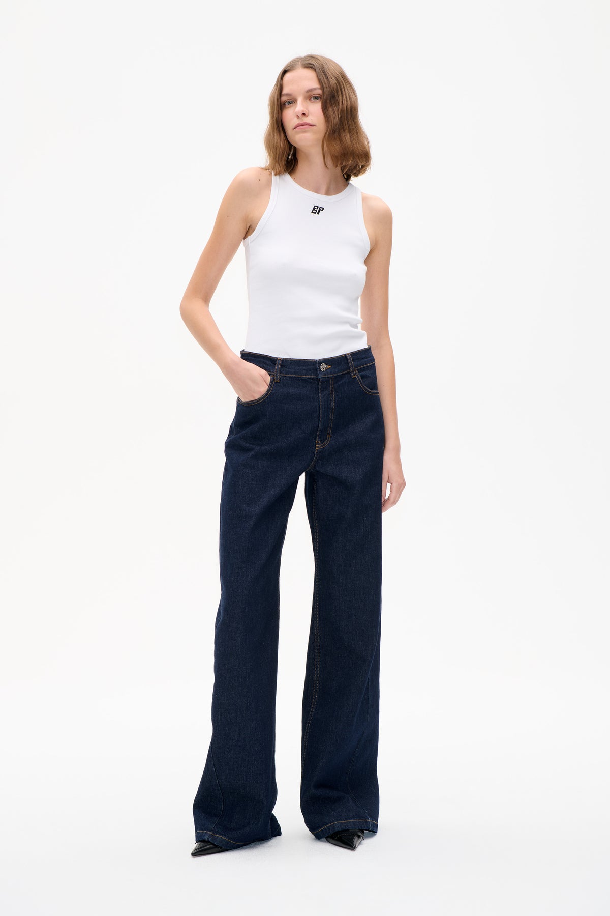 Model wearing a white ribbed high-neck tank top with a small chest logo and dark wide-leg jeans, front view.