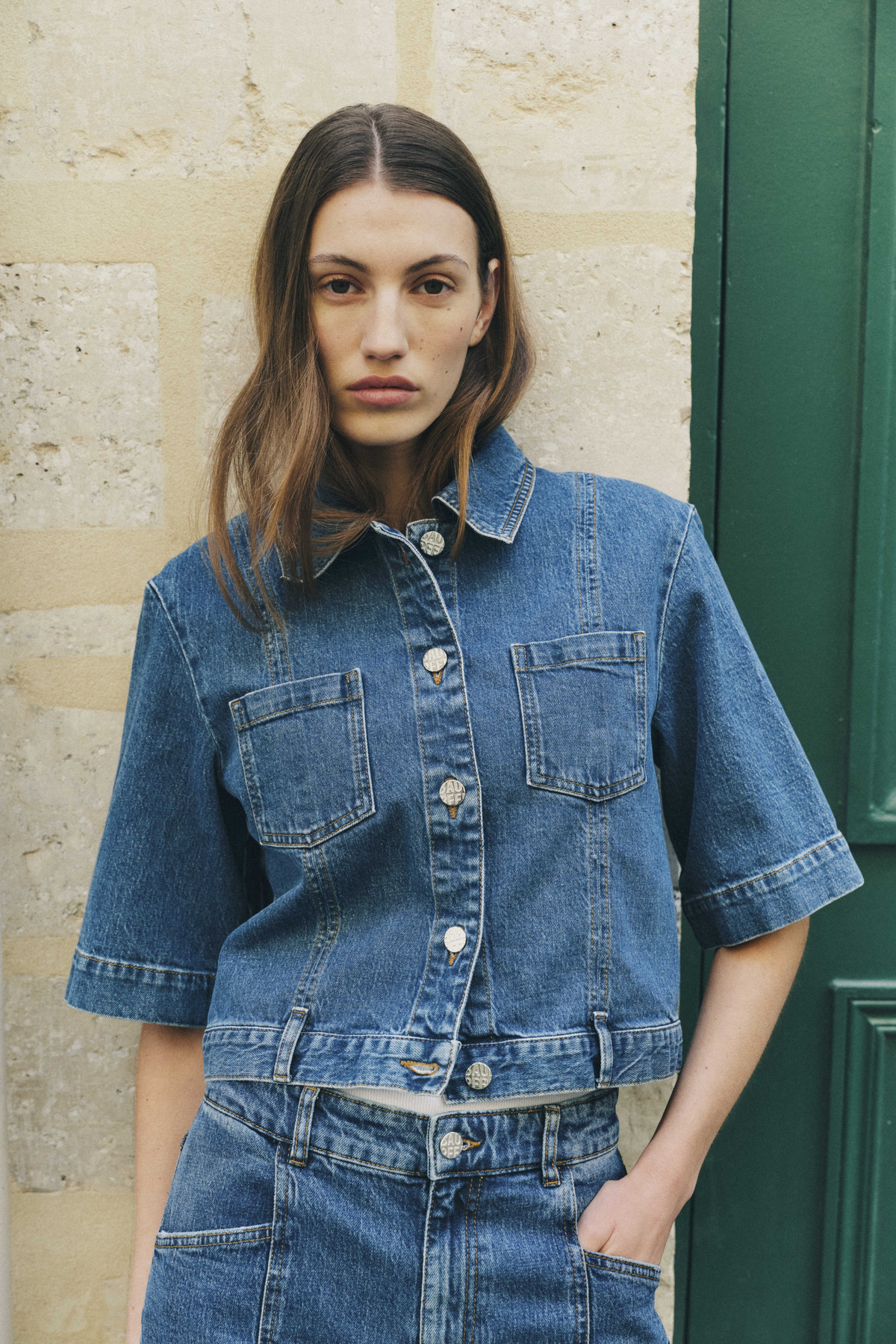 Model wearing a blue denim jacket with short wide sleeves, front buttons, two chest patch pockets and matching jeans