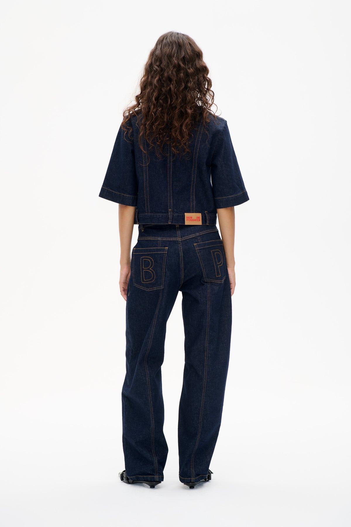 Vue de dos d'un mannequin portant une veste en denim indigo foncé et un jean à jambes larges, avec surpiqûres contrastantes et lettrages en contour sur les poches arrière.