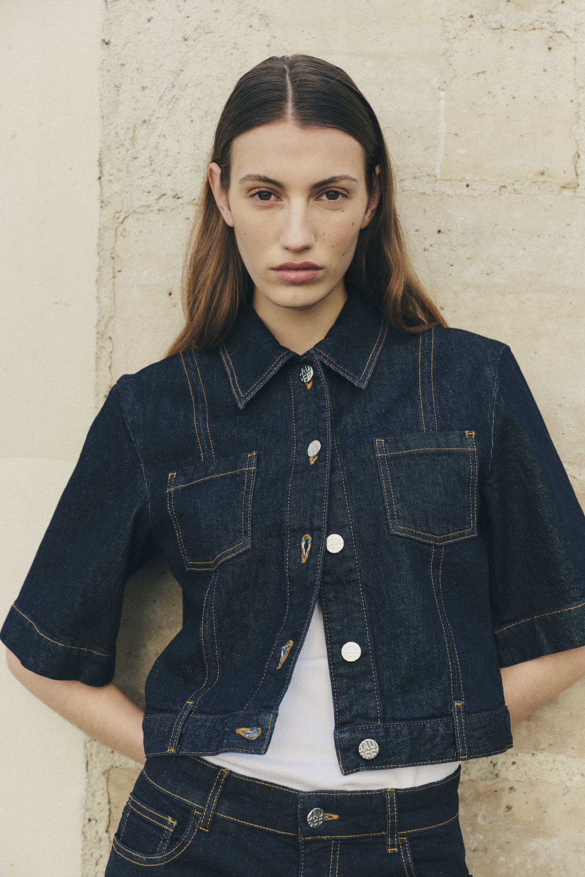 Mannequin portant une veste en denim indigo foncé à surpiqûres contrastées, col pointu, poches plaquées et boutons métalliques, portée avec un jean assorti.