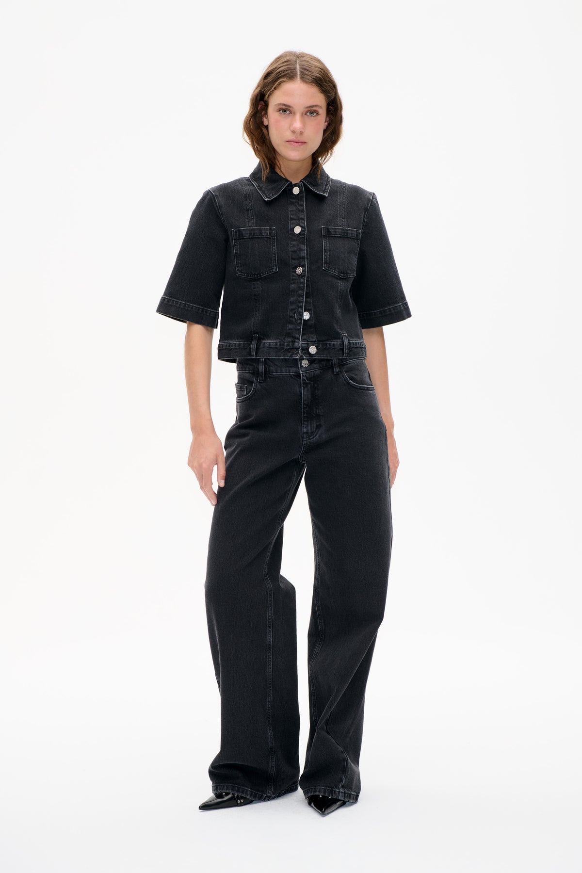 Front view of a model wearing a black denim shirt jacket with short wide sleeves, two chest pockets and matching wide-leg jeans.