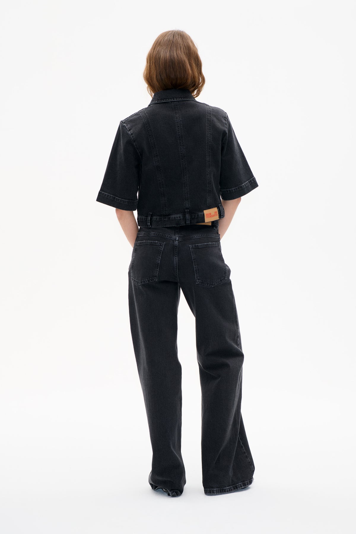 Rear view of a model wearing a black denim short-sleeved collared jacket and wide-leg jeans, with back pockets and a tan waistband patch.
