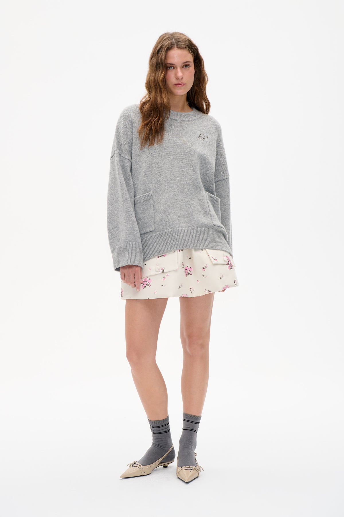 Model wearing a light grey crew-neck sweater with a small chest emblem and large front pocket, paired with a cream floral mini skirt.