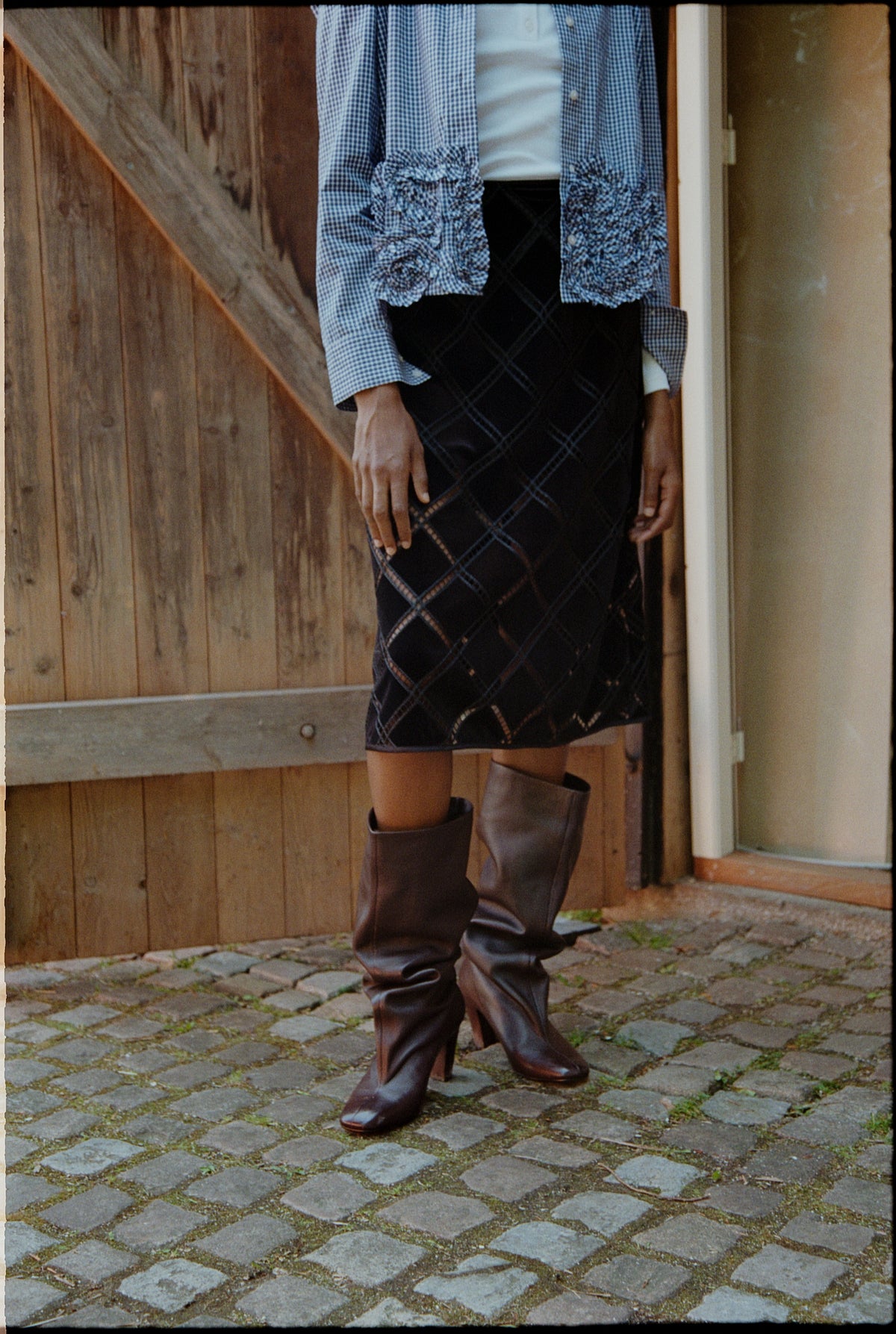 Bijgesneden campagnebeeld van een model dat een open blauw-witte gingham-blouse met ruches aan de manchetten, een zwarte geruite rok en hoge bruine laarzen draagt.
