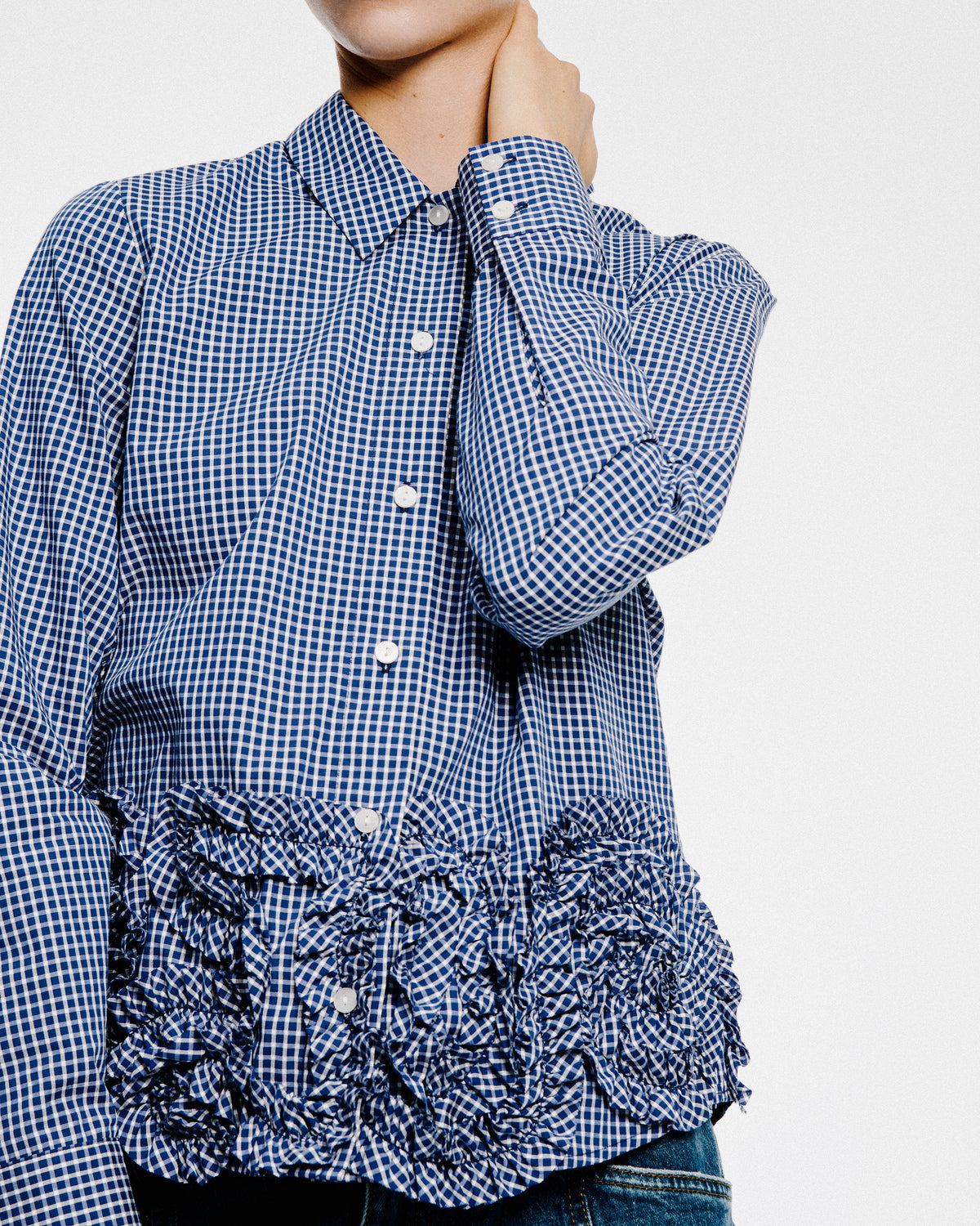 Close-up van de voorkant van een blauw-witte ruitjesblouse met knoopsluiting, puntkraag, lange mouwen en gerimpelde ruches.