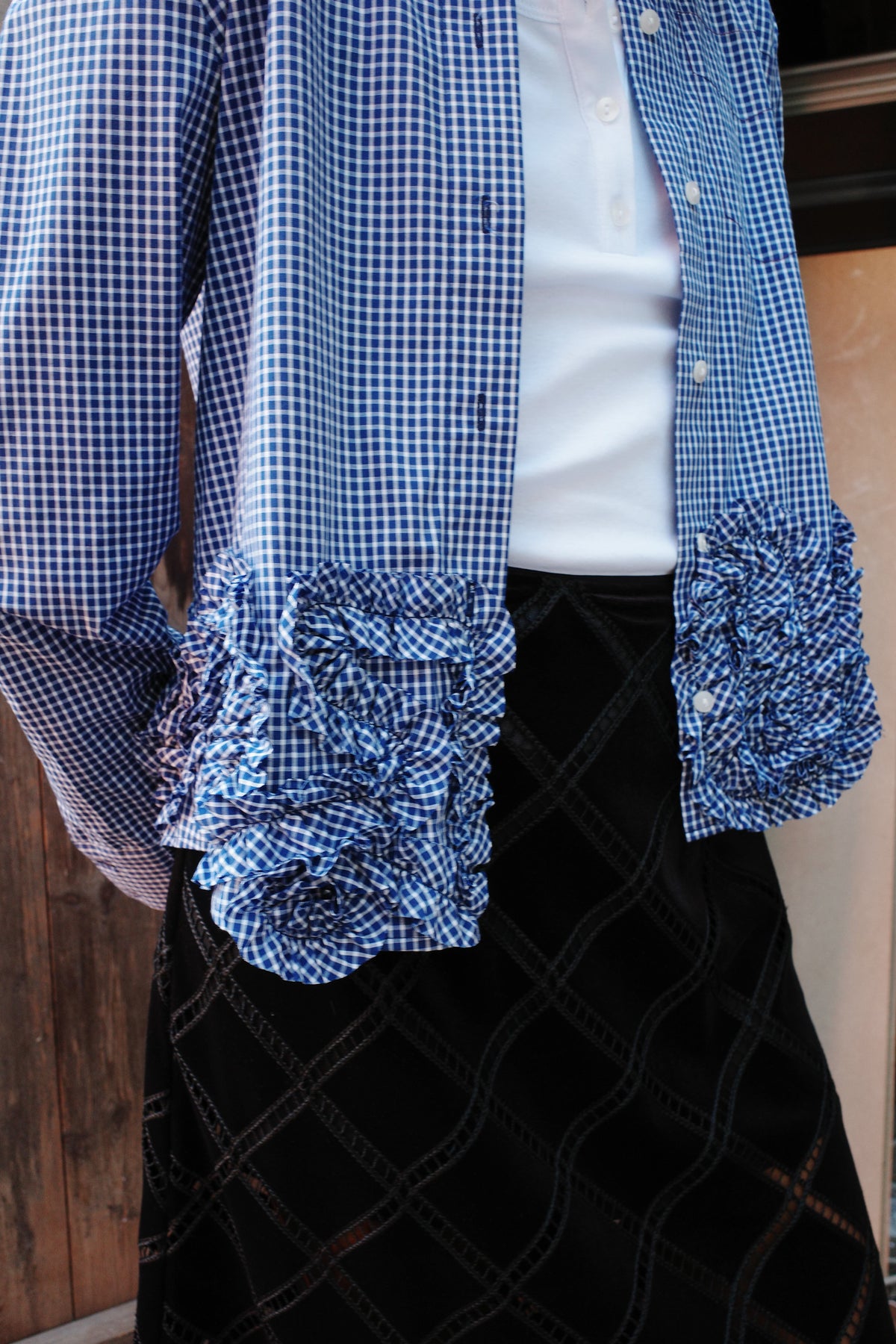Krap bijgesneden campagnebeeld van blauw-wit geruite blouses en kleding met ruches, met een witte blouse met knopen aan de voorkant en zwarte kleding zichtbaar.