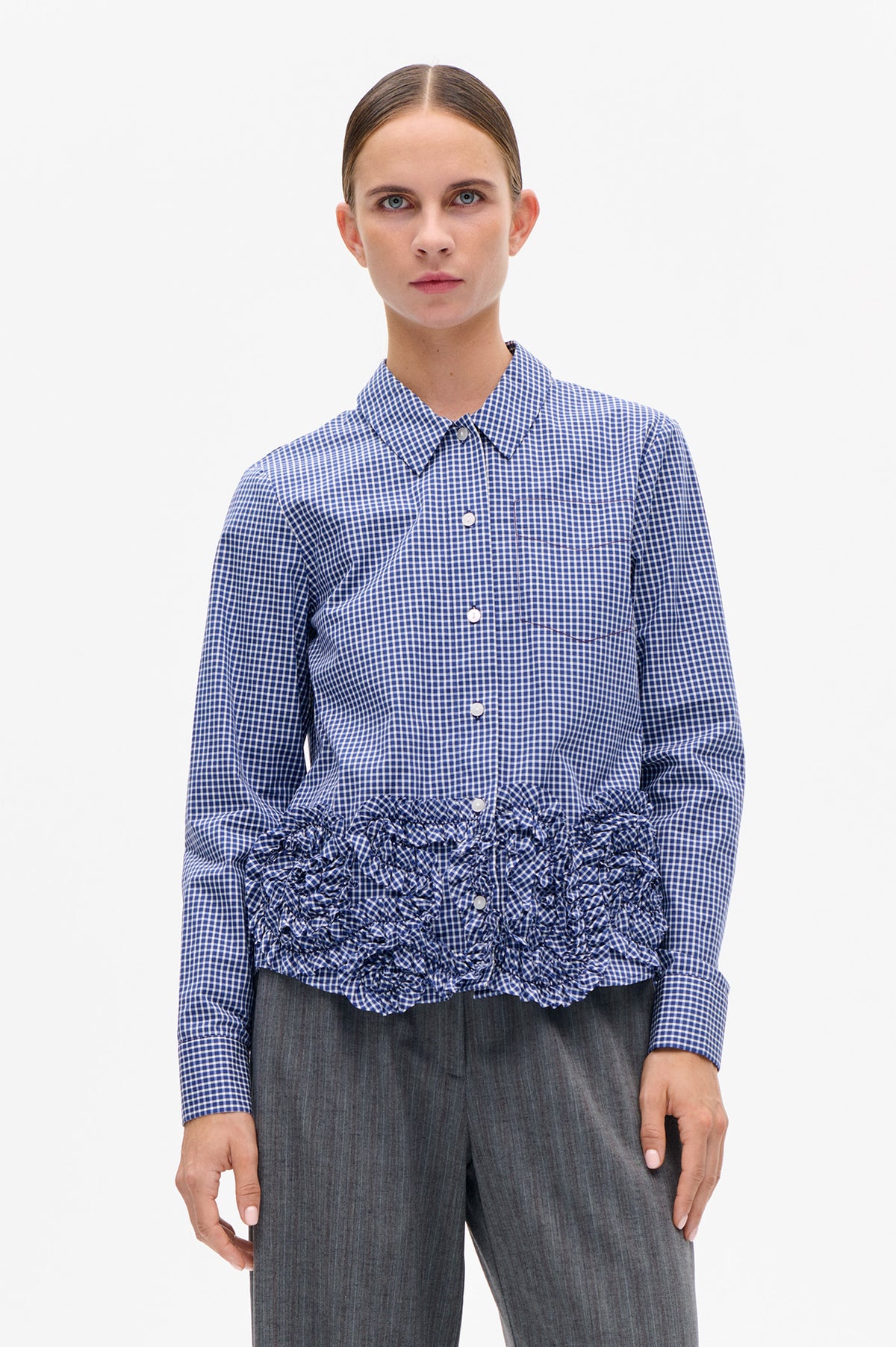 Front view of a blue-and-white gingham button-front shirt with a pointed collar, chest pocket and gathered waist, paired with gray trousers.