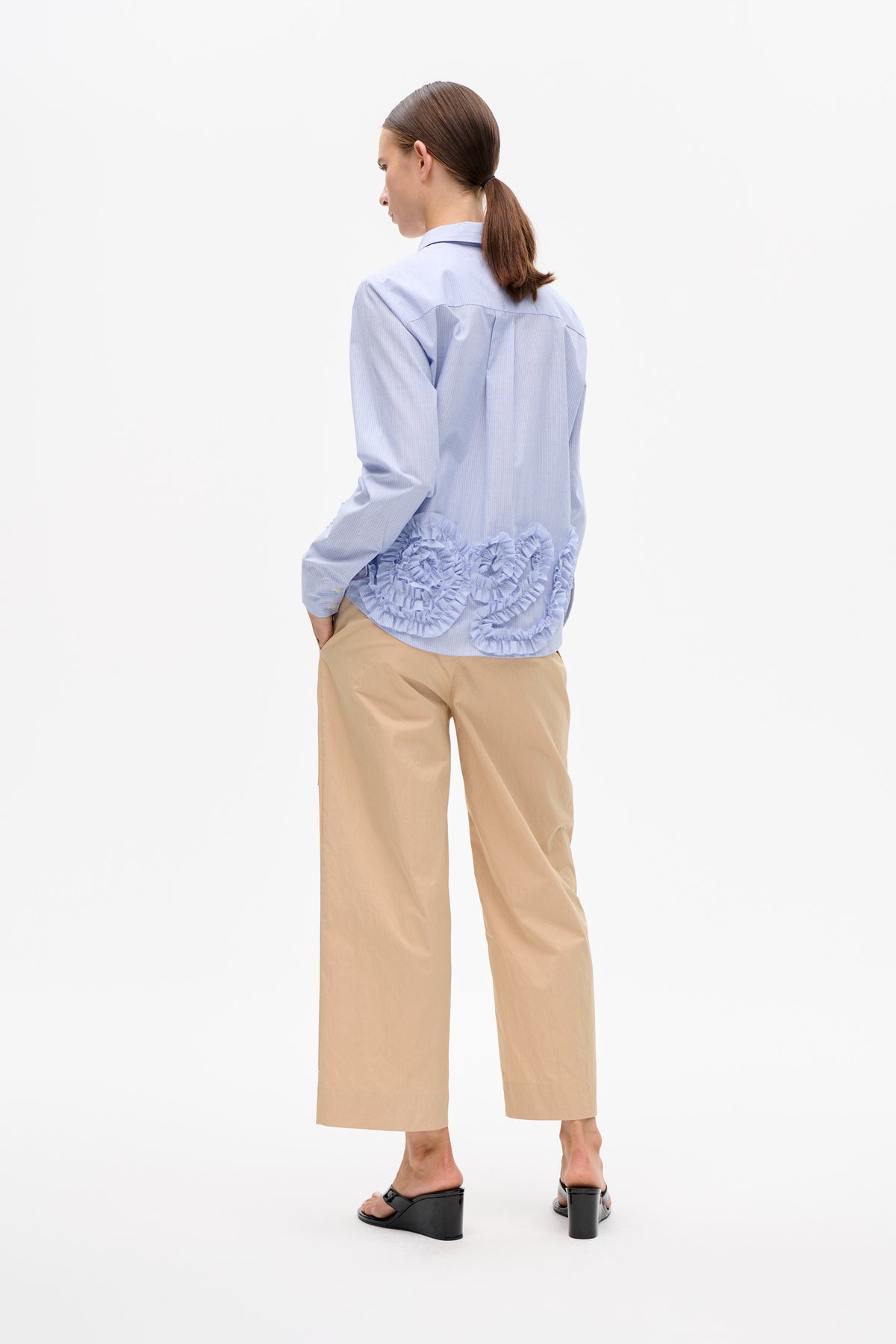 Rear view of a model wearing a light blue pinstriped button-up shirt with layered ruffle trim and beige wide-leg trousers.