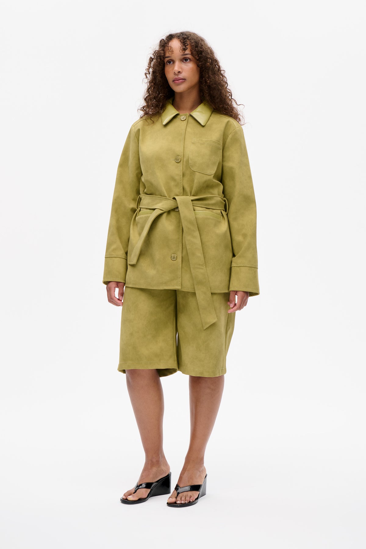 Model wearing an olive-green belted button-front jacket with a chest pocket and matching knee-length shorts, shown from the front.