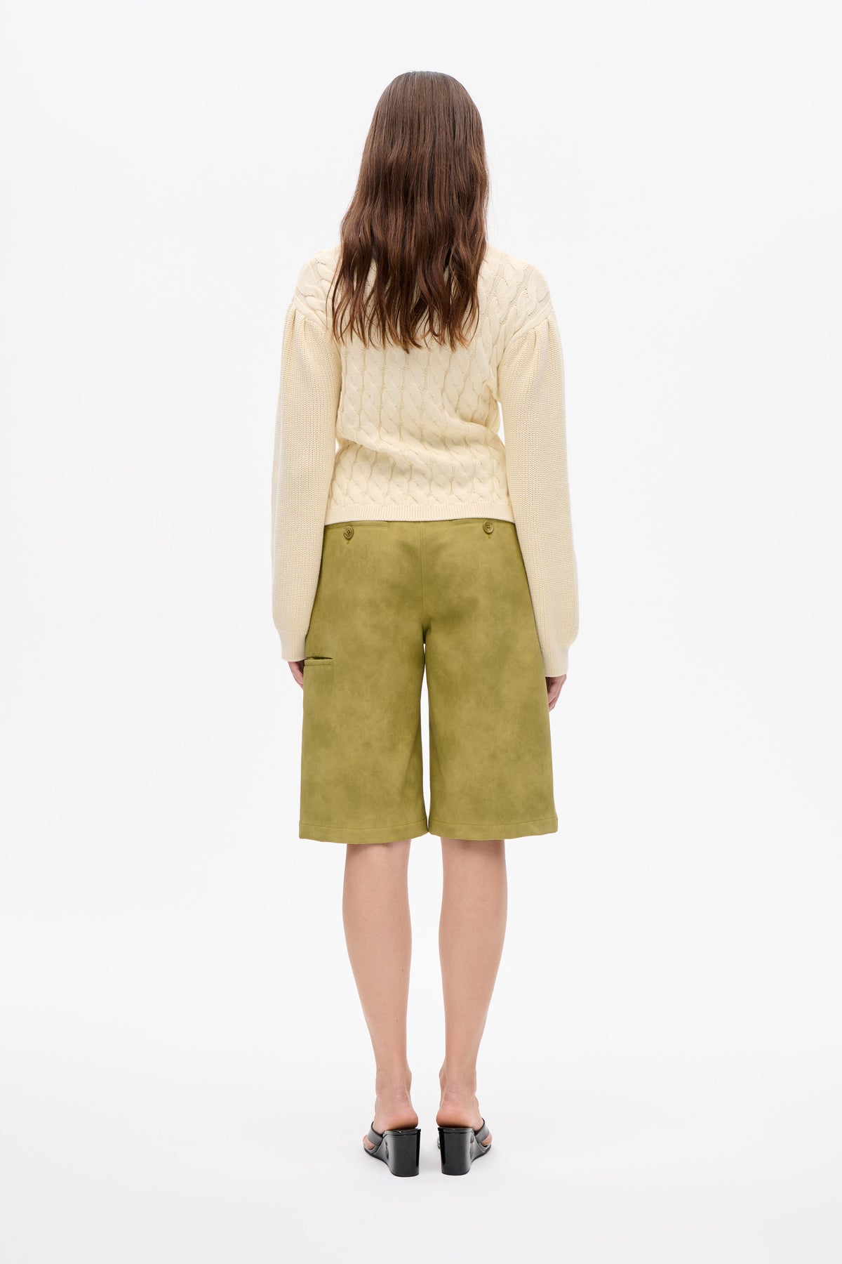 Rückenansicht eines Models, das einen elfenbeinfarbenen Zopfstrickpullover und olivgrüne, knielange Shorts mit Knöpfen im hinteren Taillenbereich trägt.