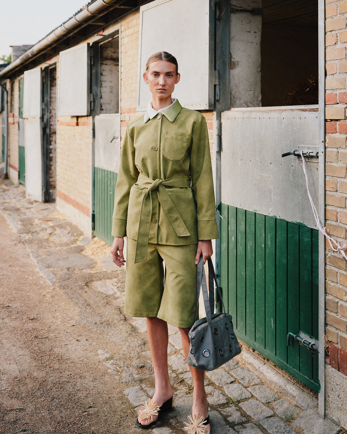 Model steht neben Stalltüren und trägt eine olivgrüne, taillierte Jacke mit Gürtel sowie passende knielange Shorts und hält eine schwarze Tasche mit Ösen.