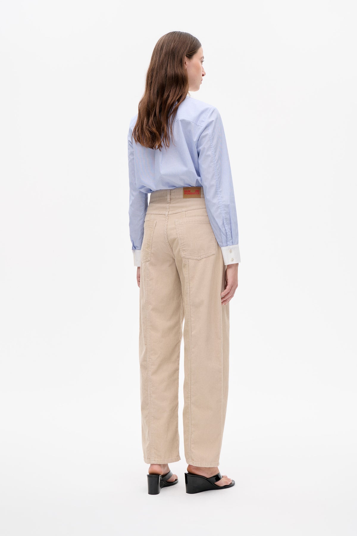 Model van achteren gezien draagt een lichtblauwe, fijngestreepte blouse met witte manchetten en een beige, wijd uitlopende broek van ribfluweel.