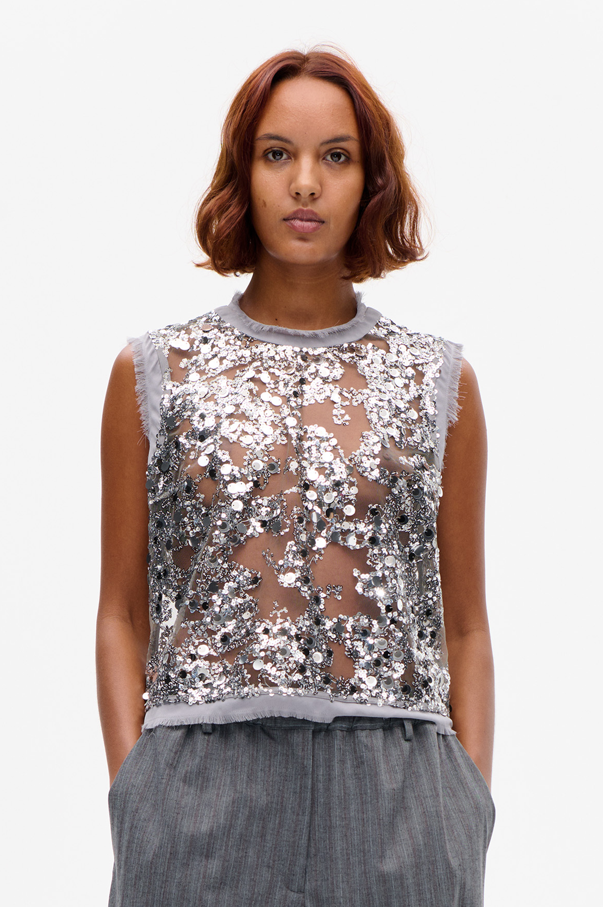 Front view of a sheer sleeveless top densely covered in silver, grey and black sequins, with a pale grey fringed neckline and edges.