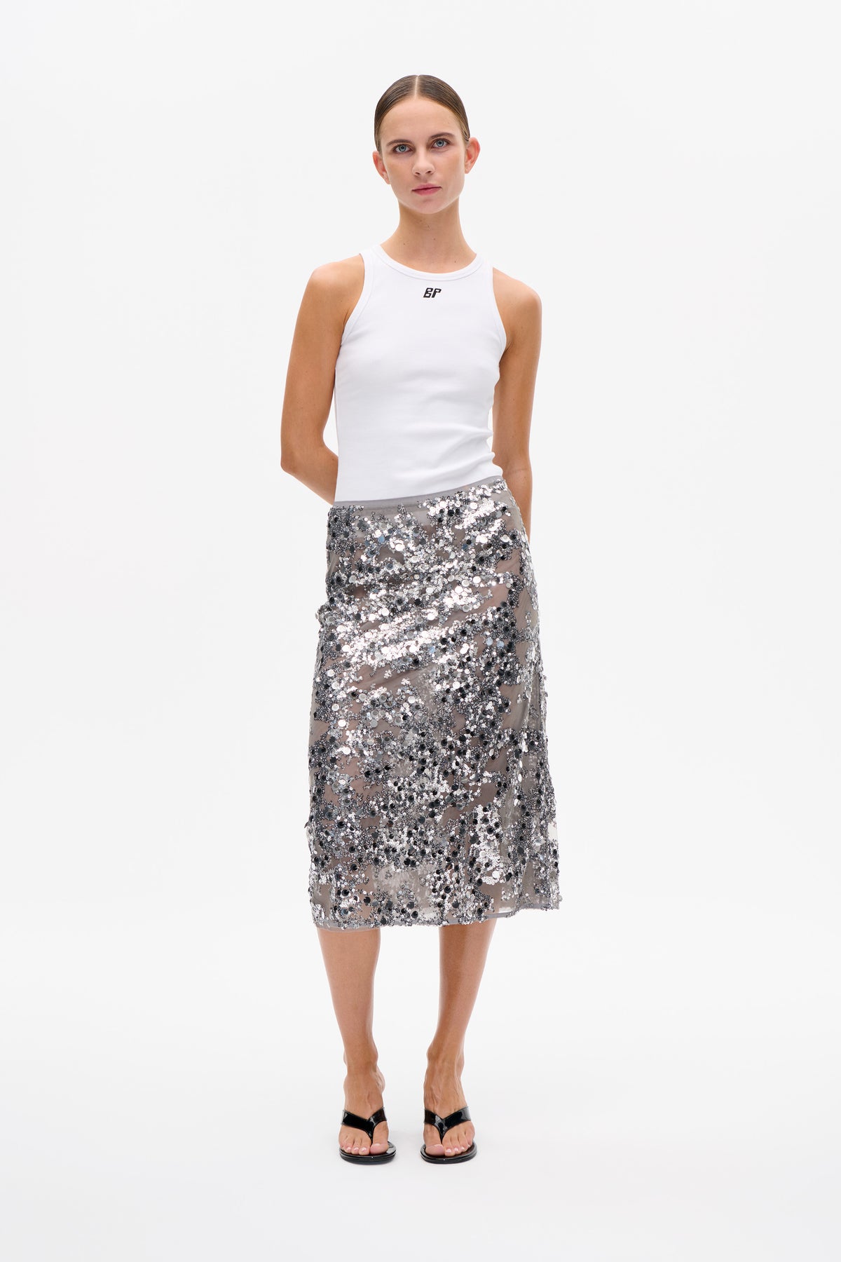 Model wearing a white sleeveless ribbed top with a small chest logo and a sheer grey knee-length skirt covered in silver and black sequins.