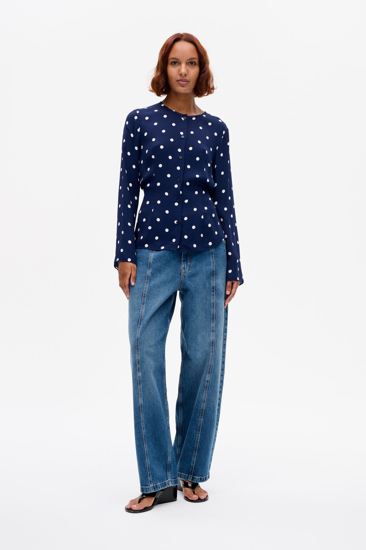 Vorderansicht eines Models, das eine marineblaue, langärmelige Bluse mit Knopfleiste und weißen Polka-Dots sowie eine blaue Jeans trägt.