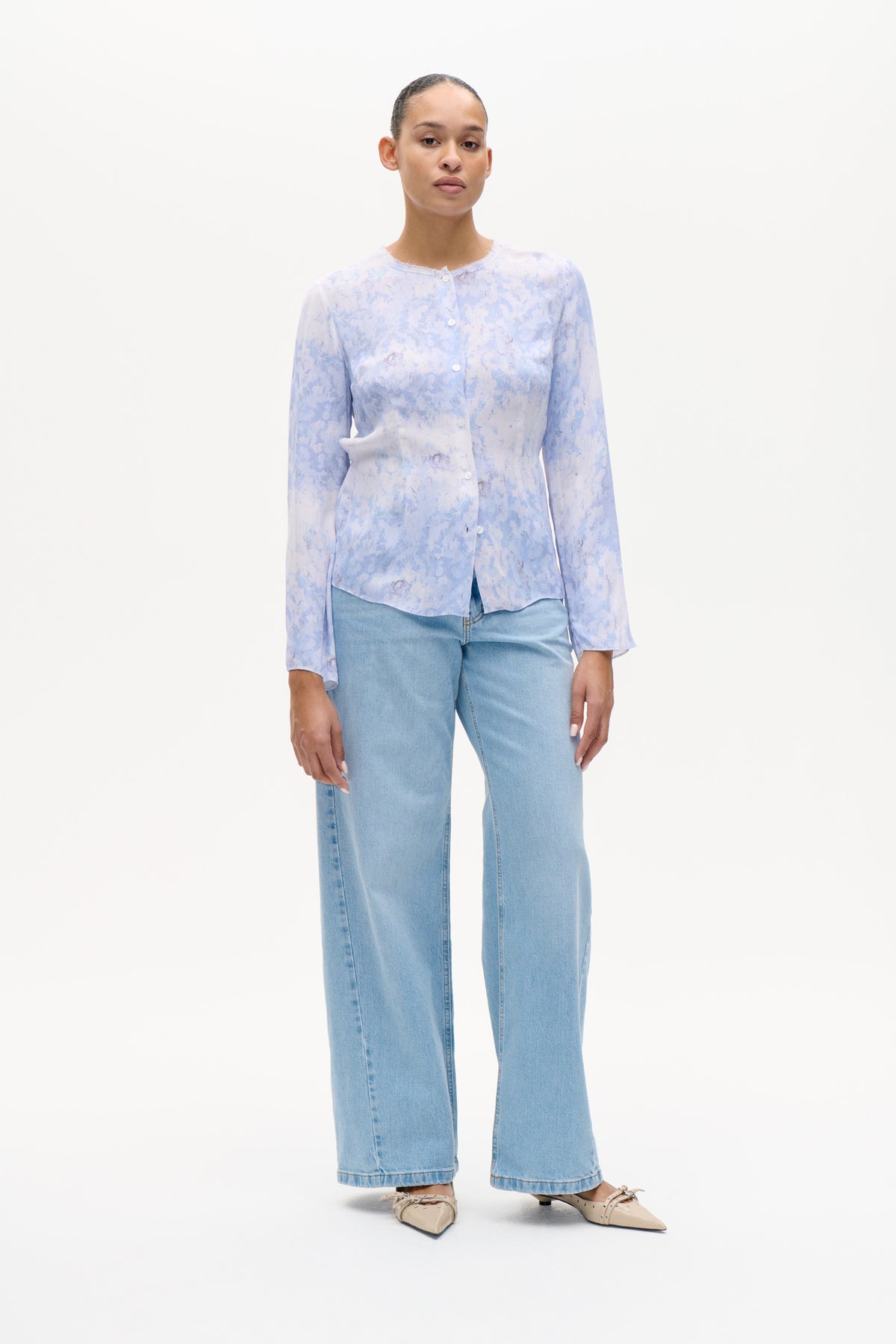 Vooraanzicht van een model dat een lichtblauwe en witte blouse met bloemenprint, knoopsluiting en lange mouwen draagt, gecombineerd met lichtgewassen jeans met wijde pijpen.