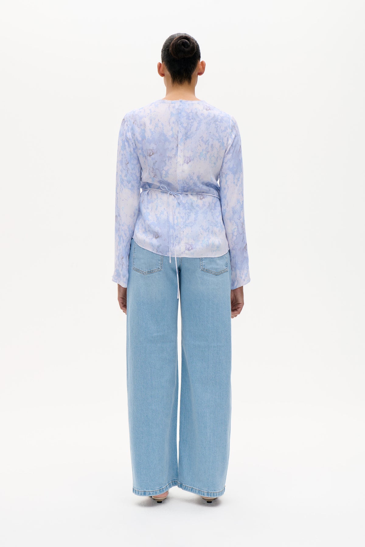 Achteraanzicht van een model dat een lichtblauwe, lavendelkleurige blouse met abstracte bloemenprint en een taillestrik draagt, gecombineerd met lichtgewassen, wijduitlopende jeans.