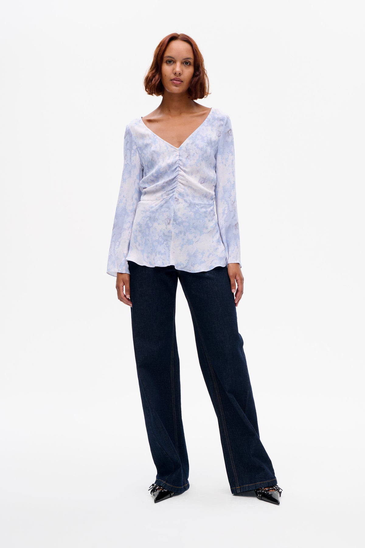 Vorderansicht eines Models, das eine hellblaue Bluse mit abstraktem Blumenmuster, V-Ausschnitt und gerafftem Vorderteil trägt, kombiniert mit dunklen, weit geschnittenen Jeans.