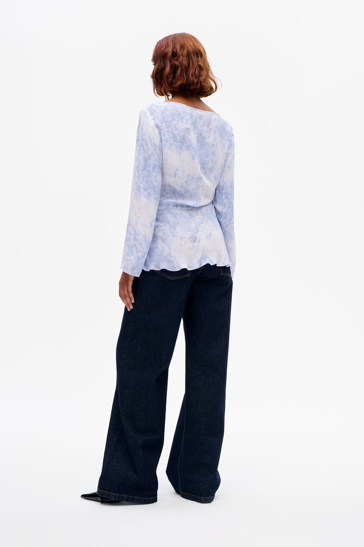 Rückenansicht eines Models, das eine hellblaue, geblümte Langarmbluse mit weitem Ausschnitt und eine dunkle, weit geschnittene Jeans trägt.