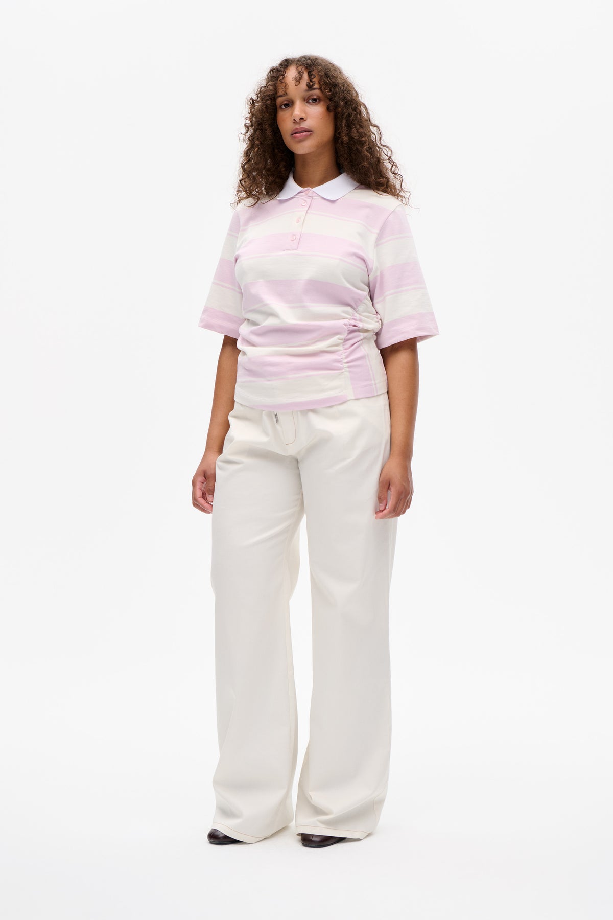 Vorderansicht eines Models in einem kurzärmligen Poloshirt mit rosa- und cremefarbenen Streifen, weißem Kragen und weiten cremefarbenen Hosen.