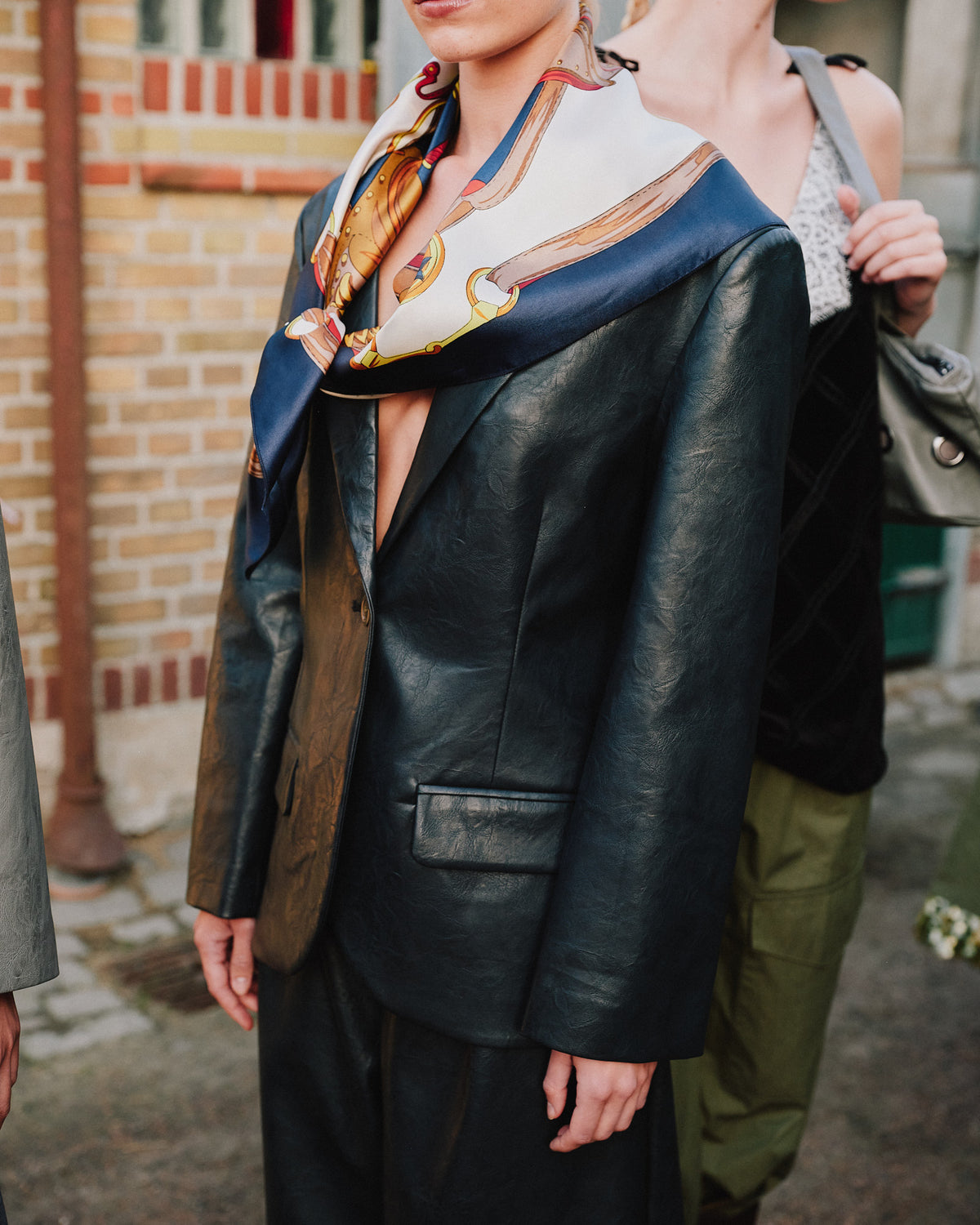 Campaign close-up of a model in a dark leather-look blazer with a cream and navy illustrated scarf; other models stand behind her.