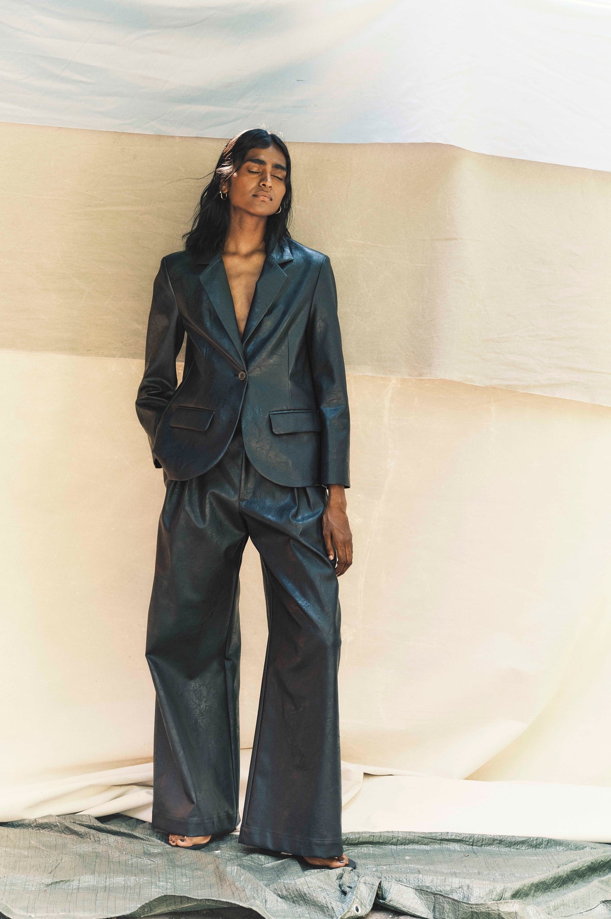 Campaign portrait of a model wearing a dark teal glossy blazer with notched lapels and wide-leg trousers against a cream backdrop.