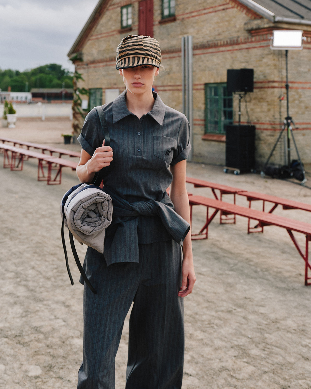 Campaign portrait of a model in a charcoal subtly striped short-sleeve button-up and wide-leg trousers, wearing a striped cap and carrying a grey backpack.