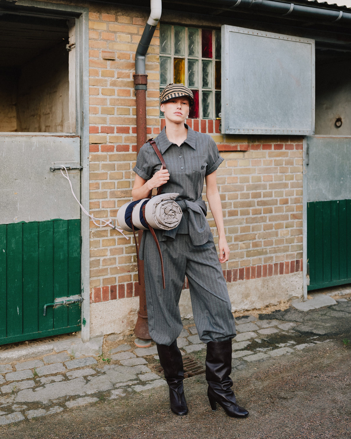 A model wears a charcoal-gray short-sleeved button-up shirt with matching wide-leg trousers and black boots, carrying rolled garments.